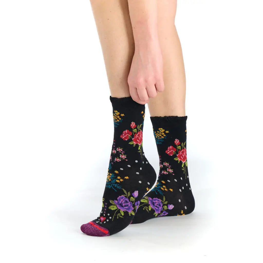 A person's legs and feet wearing black crew socks with a colorful floral pattern of red, purple, and yellow flowers, shown against a plain white background.