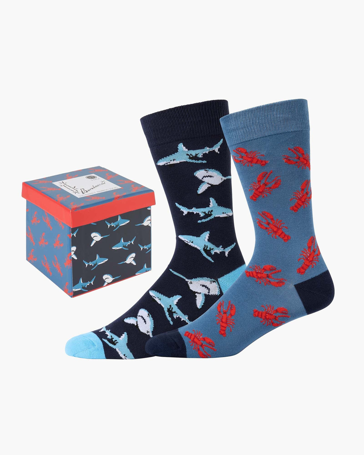 A pair of novelty crew socks with a split design: one sock is navy blue with light blue and white sharks, and the other is light blue with red lobster illustrations. A matching patterned gift box is shown next to them on a light gray background.
