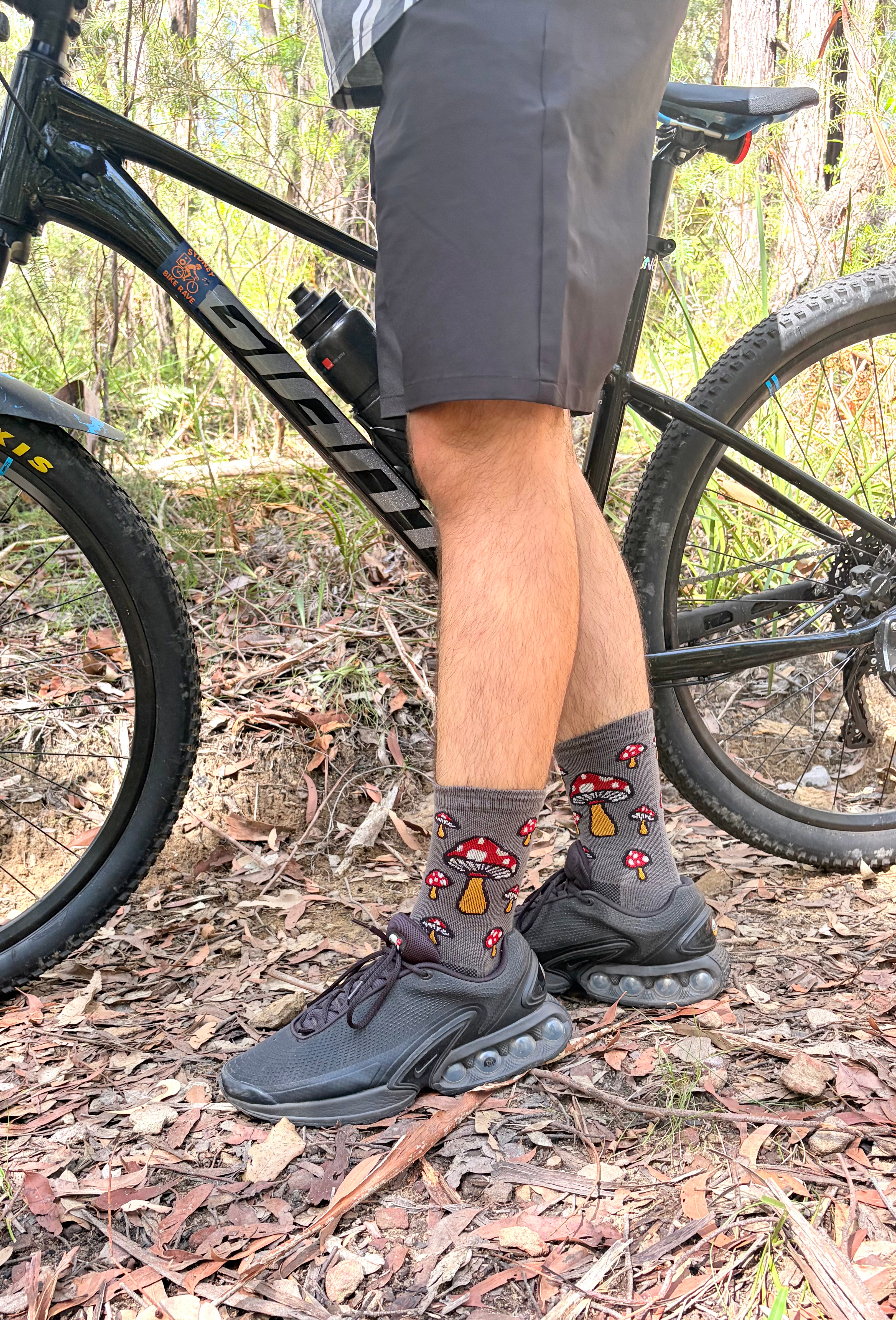 Person wearing black sneakers and sockguy socks with Mushroom design on a bike in a natural setting