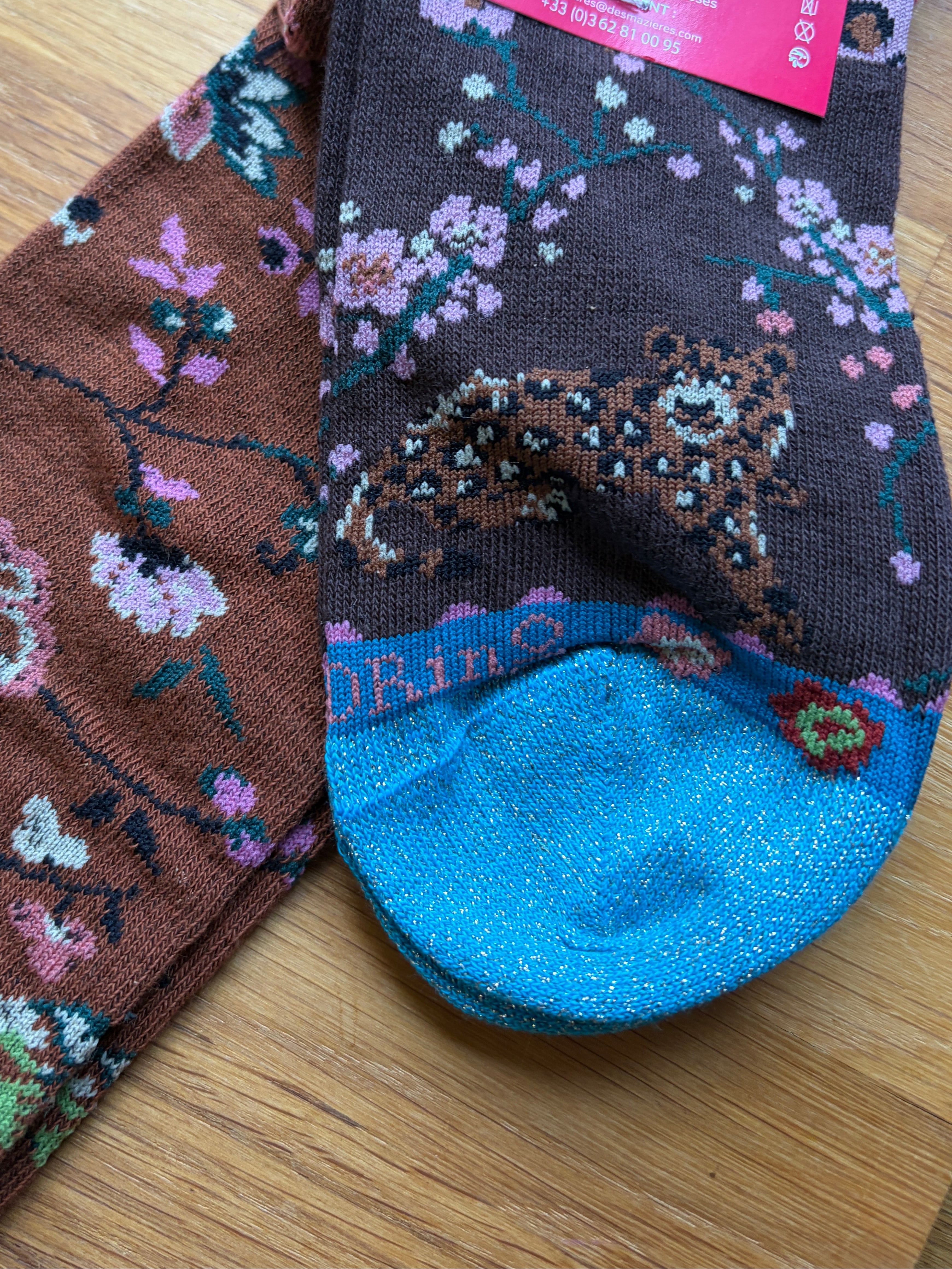 A pair of patterned crew socks laid flat on a wooden surface. One sock is dark brown with a floral branch design in pink, blue, green, and white, and the other is dark gray with a leopard print and pink floral accents.