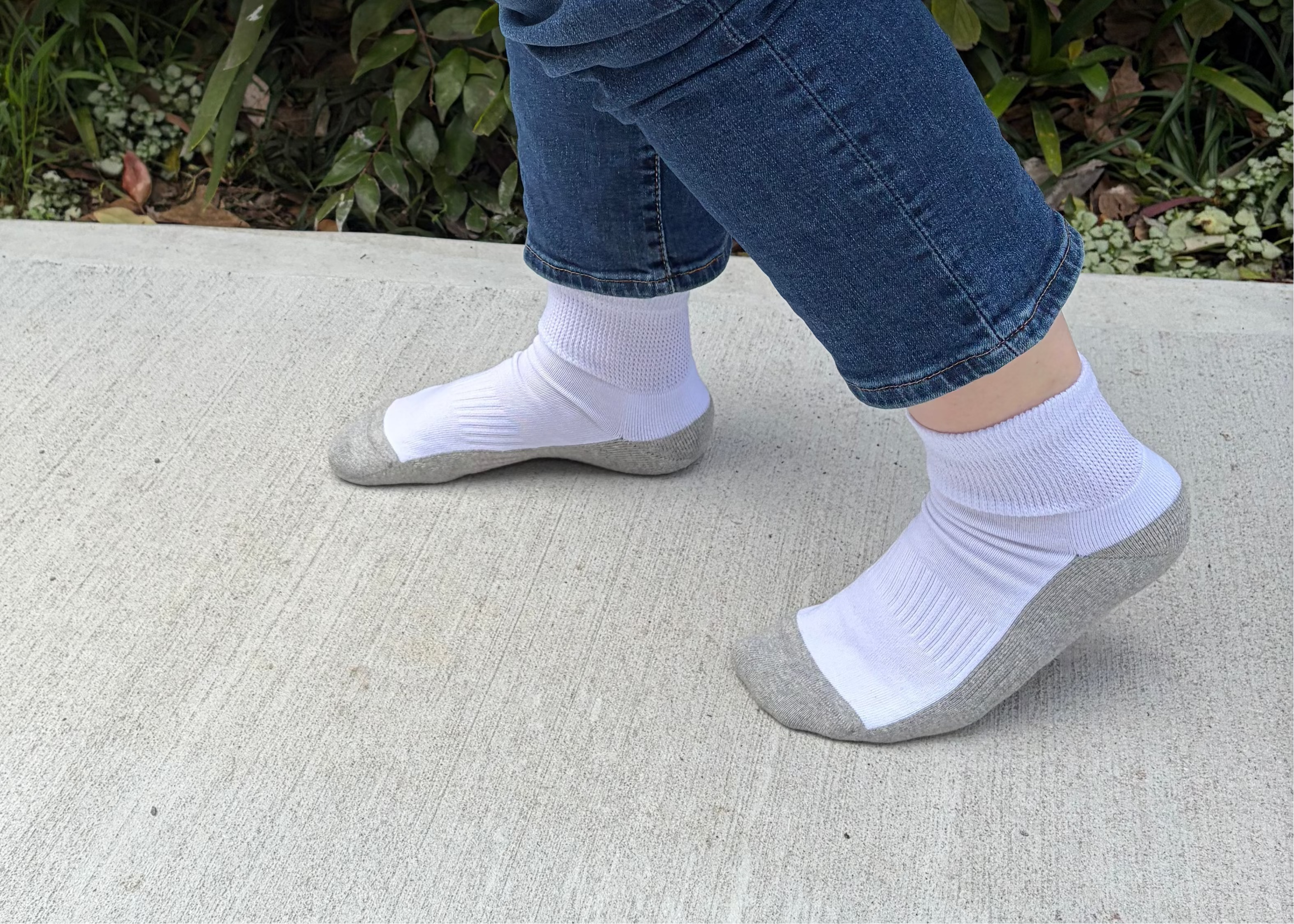 White athletic socks with gray toe and heel sections worn by a person in blue jeans, standing on a concrete surface with green foliage in the background.