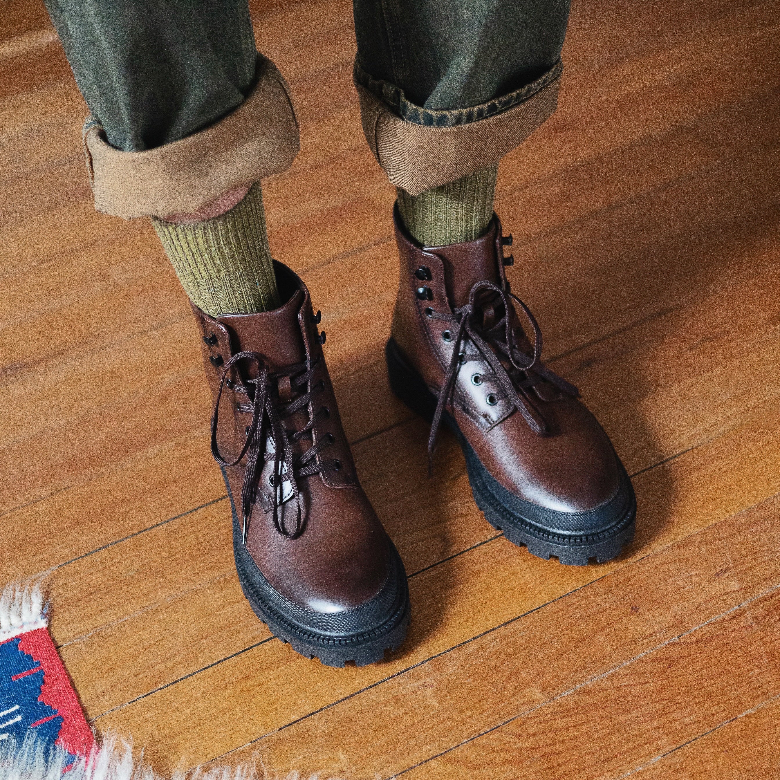 Brown leather boots worn with green pants and socks on a wooden floor.