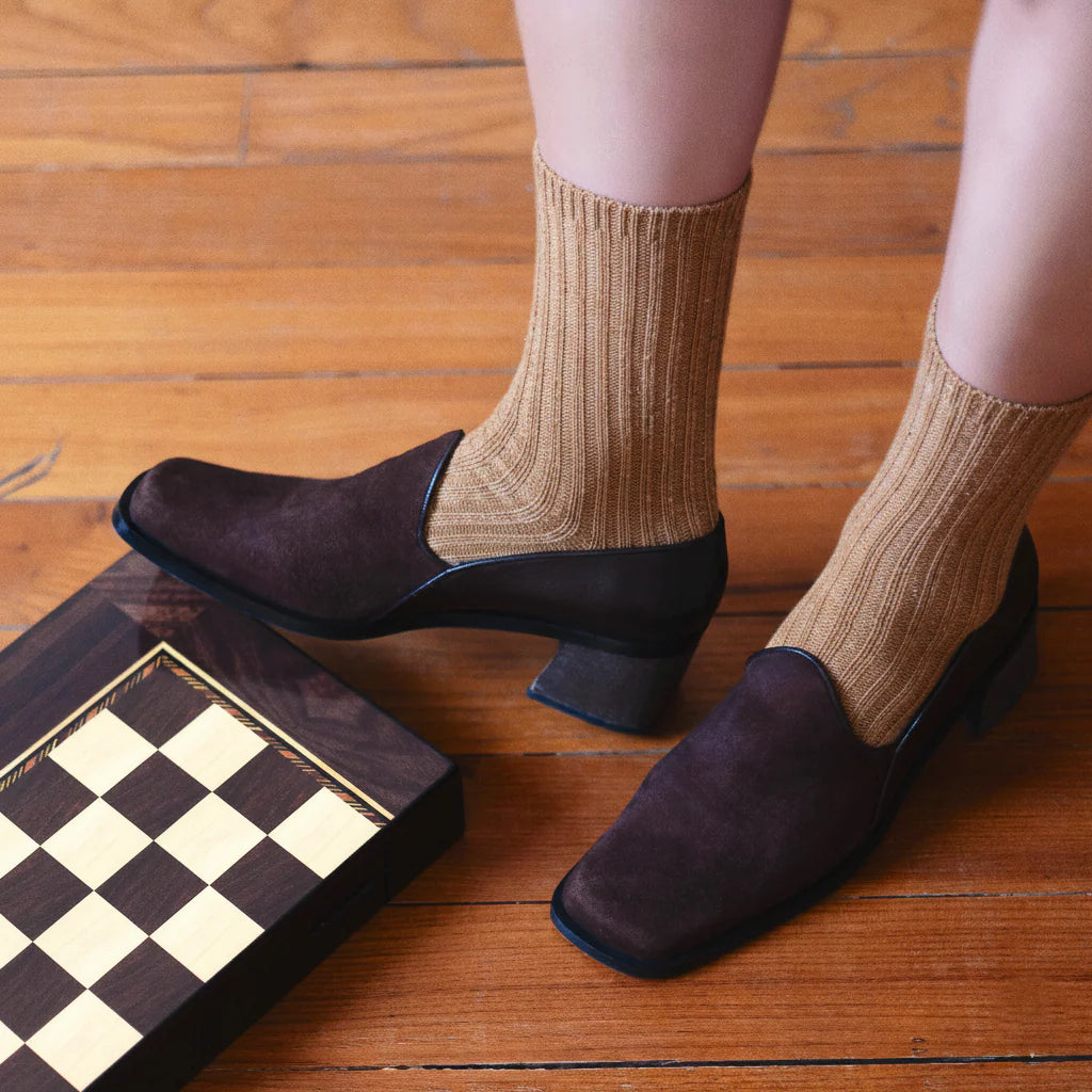 Person wearing brown shoes and beige socks on a wooden floor with a chessboard.