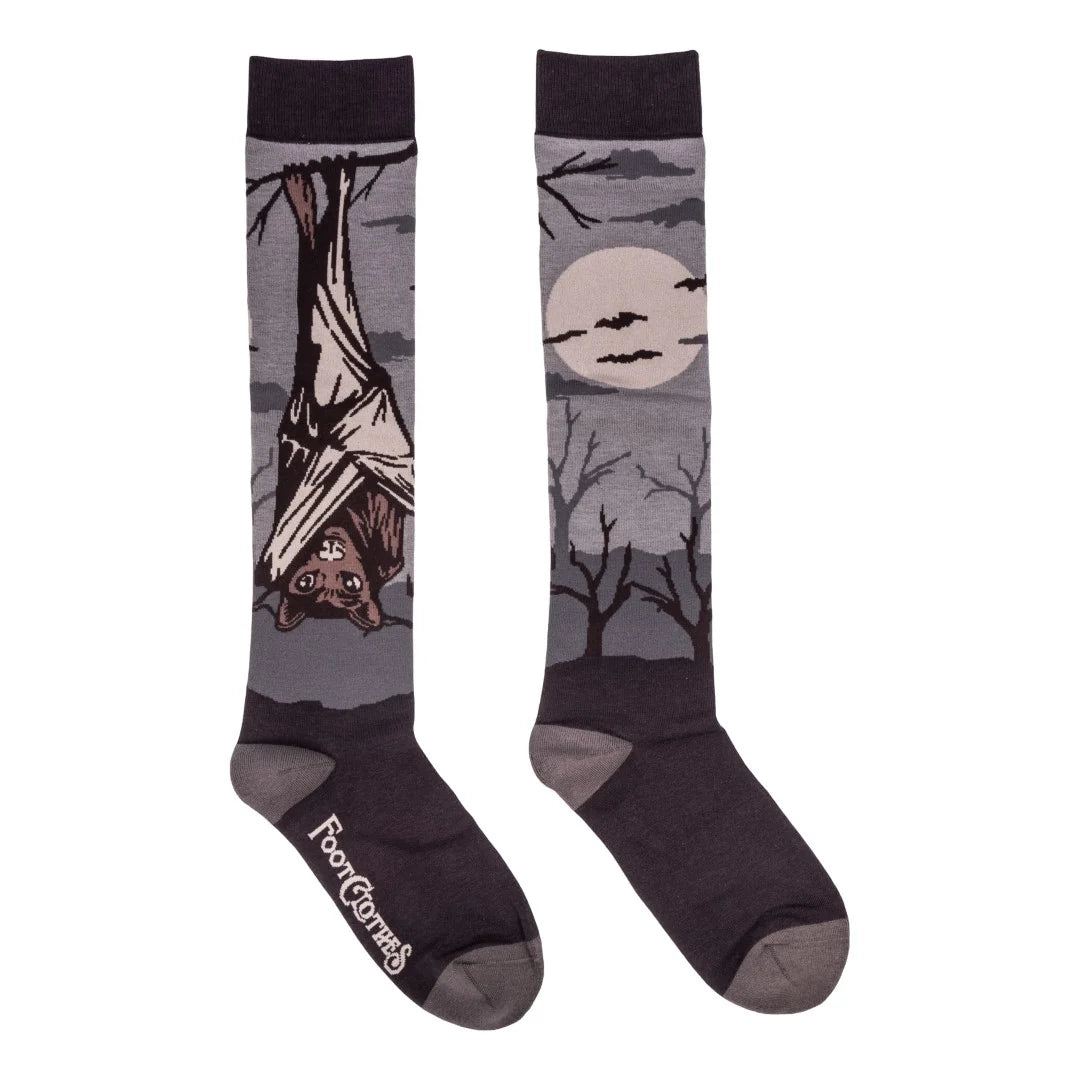 A pair of crew socks with a spooky Halloween theme, featuring a bat hanging from a tree branch on one sock and a full moon, bats, and bare trees on the other, in dark gray and black with a light gray sole.