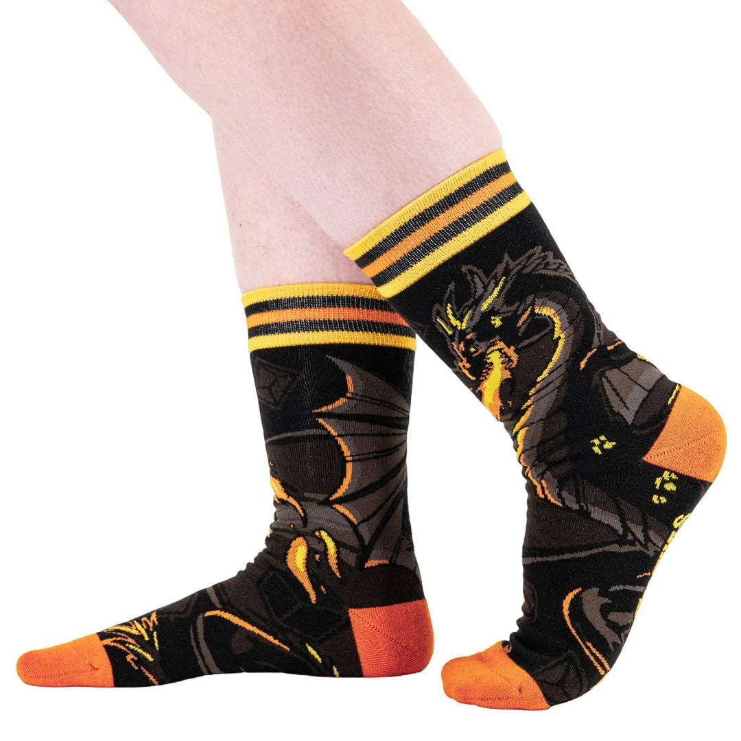 A pair of crew socks worn on feet, featuring a black background with a detailed yellow and orange dragon design, yellow and black striped cuffs, and bright orange toes and heels.