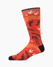 A single crew sock with a vibrant red and orange background featuring an Aboriginal-inspired pattern of black footprints, white bird silhouettes, and orange geometric lines, shown on a plain white background.