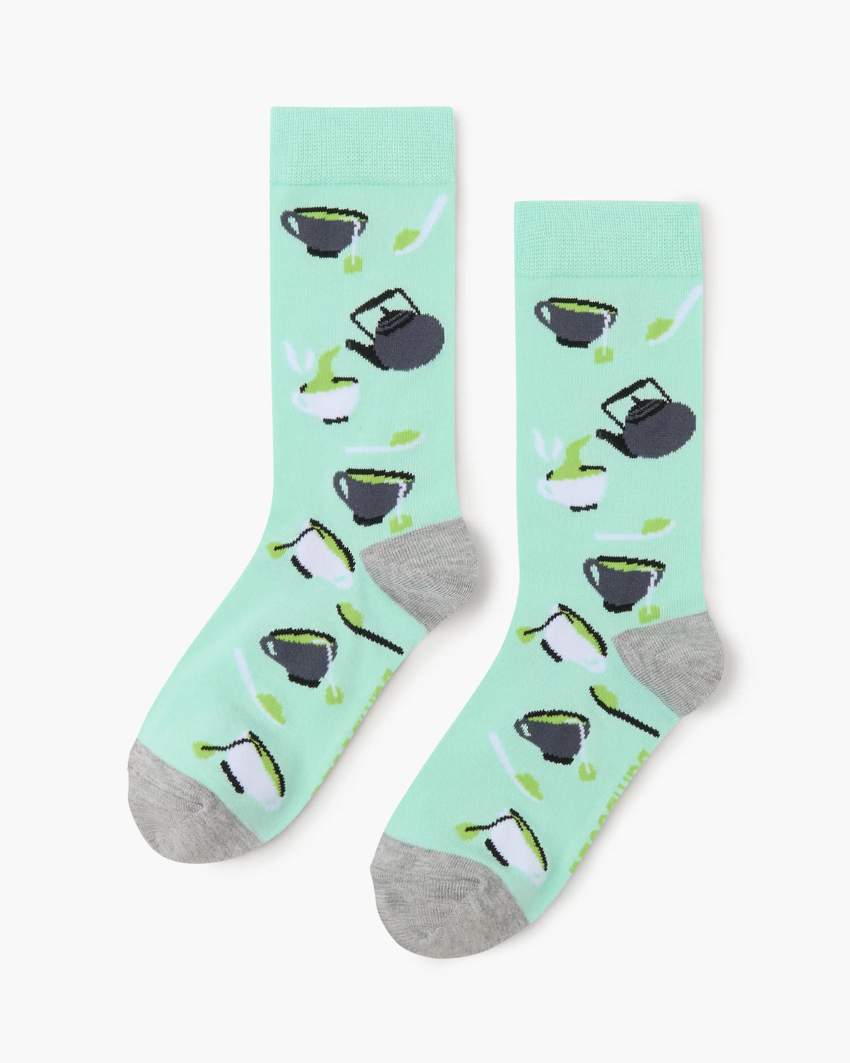 A pair of mint green crew socks with a pattern of black teapots pouring green tea into cups, featuring gray toes and heels, displayed on a light gray background.