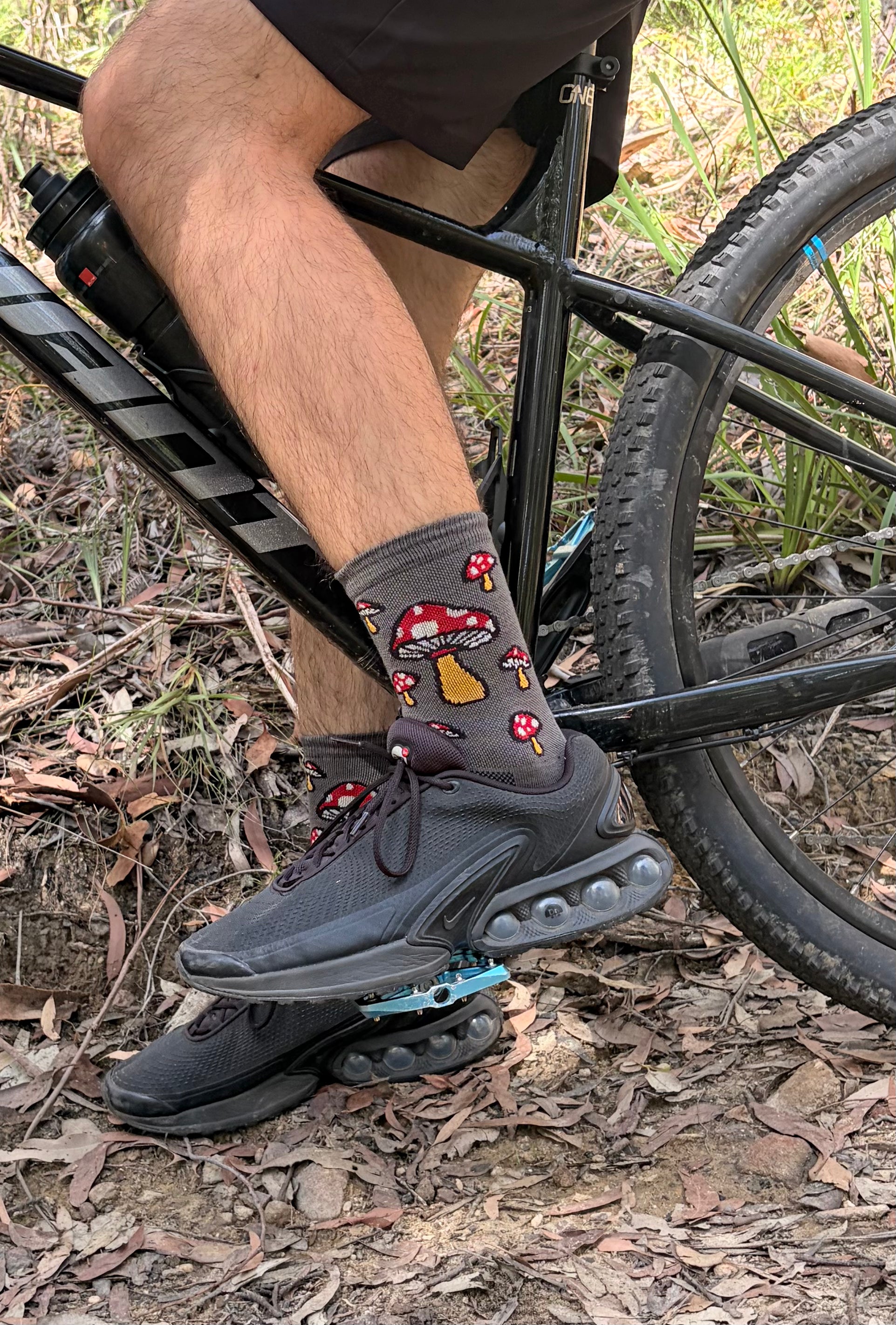 Person wearing black sneakers and sockguy socks with Mushroom design on a bike in a natural setting
