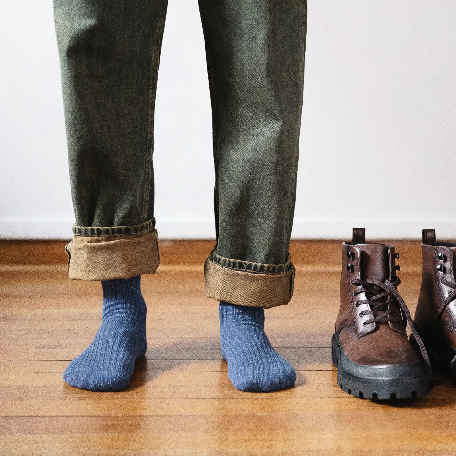 Person wearing blue socks with rolled-up green pants, standing next to a pair of brown boots on a wooden floor.