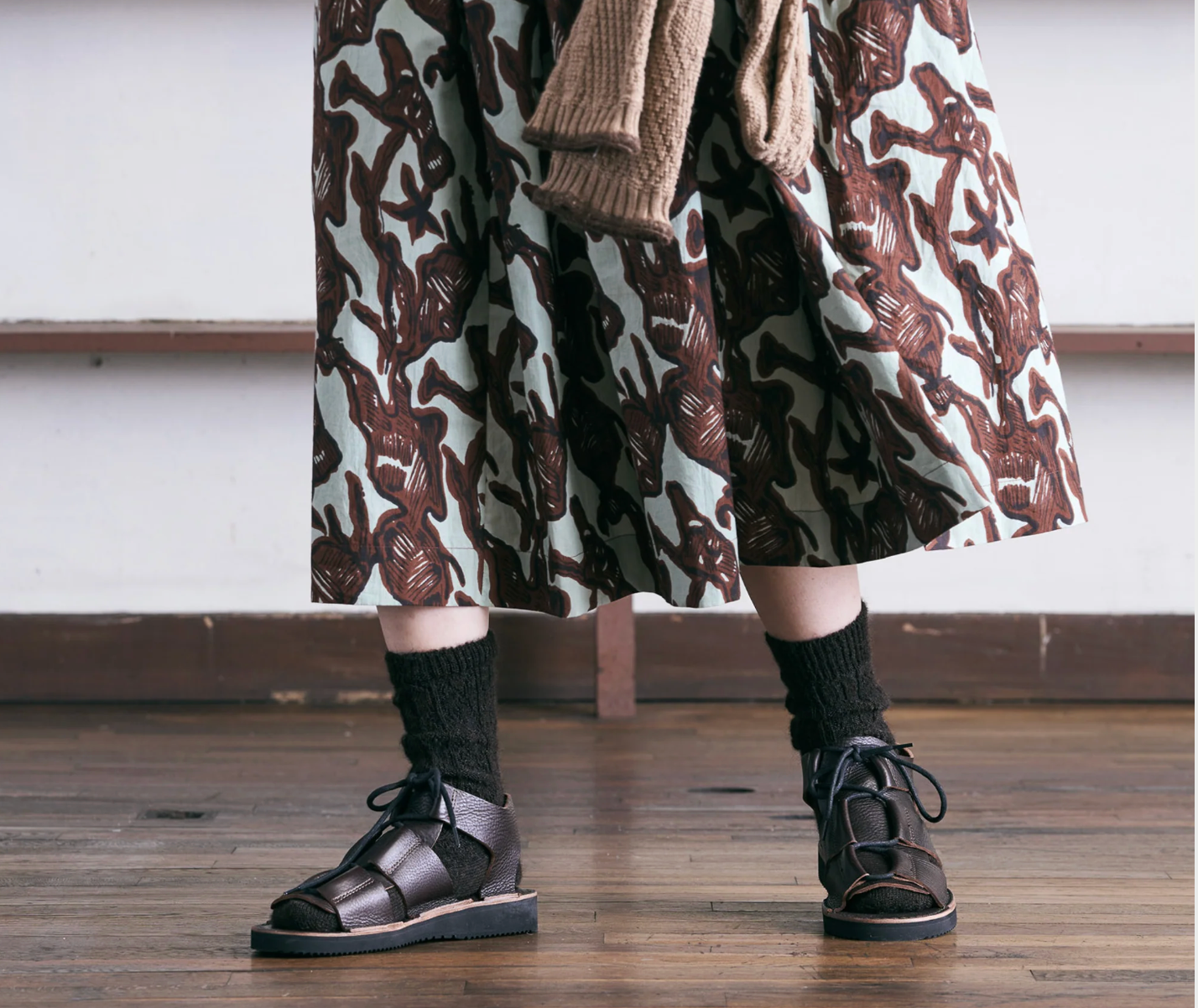 Person wearing a patterned skirt, cardigan, and sandals on a wooden floor.