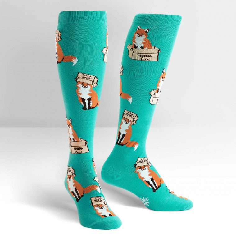 Light green knee high socks with foxes all over.  Some sit in a box, others wear the box on their heads. The box has the word box crossed out, and the word fox written underneath - The Sockery