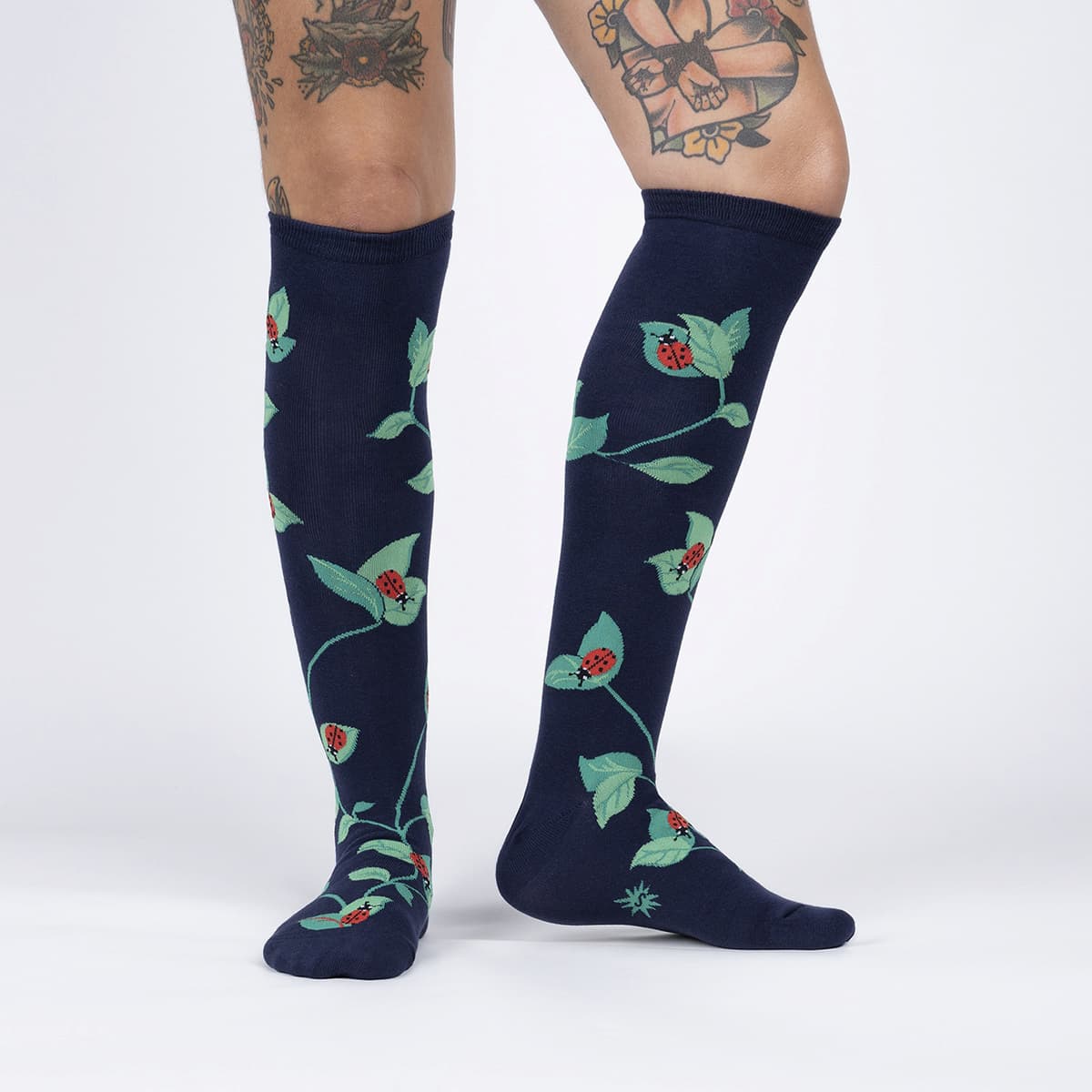 Person with tattoos wearing navy blue knee high socks with green leaves with red ladybugs on them all over - The Sockery