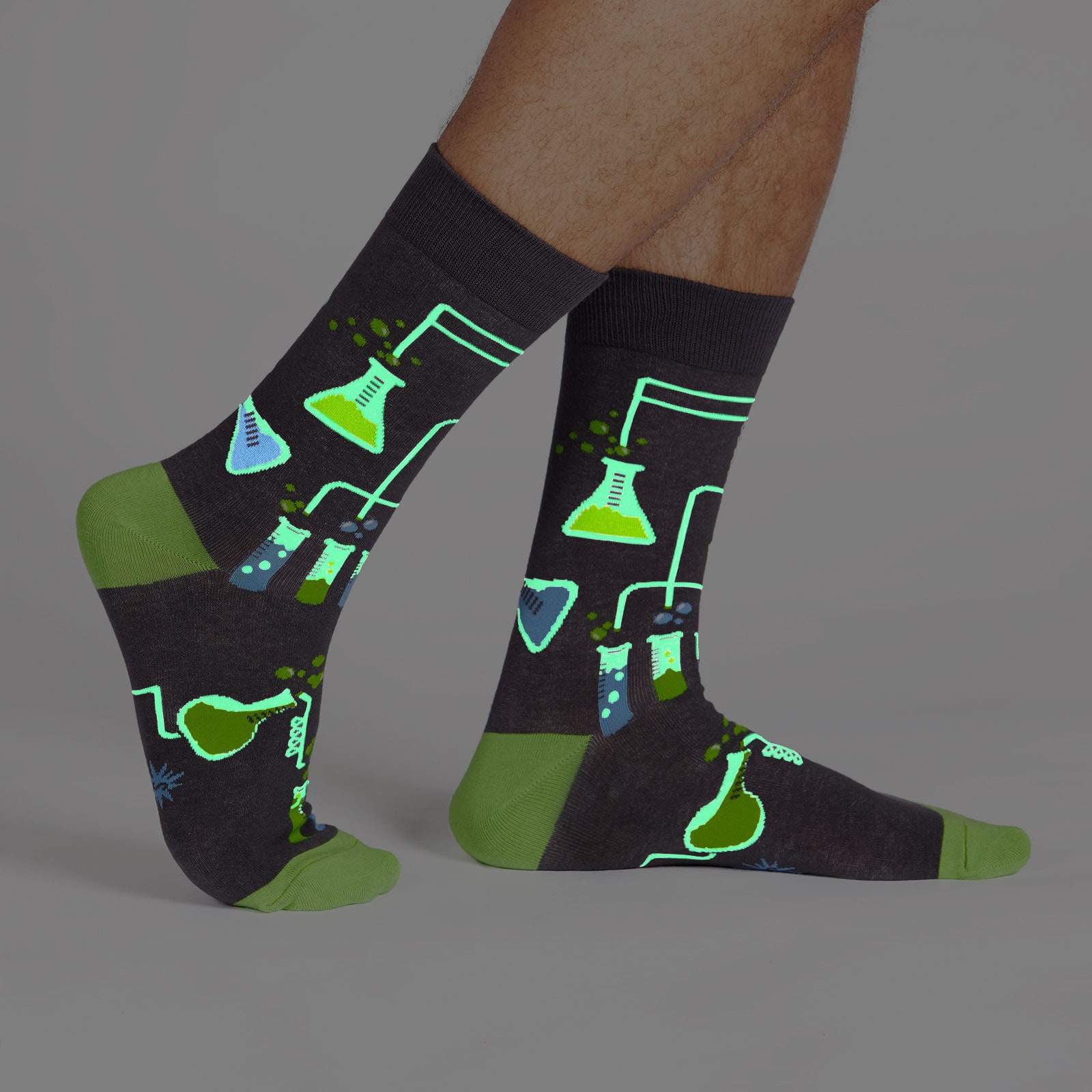 Featuring all things found in a good science lab - beakers, test tubes, glowing liquids, condensation flasks on a  steel grey mens crew sock. Image is taken in the dark, and the lab equipment is glowing - The Sockery